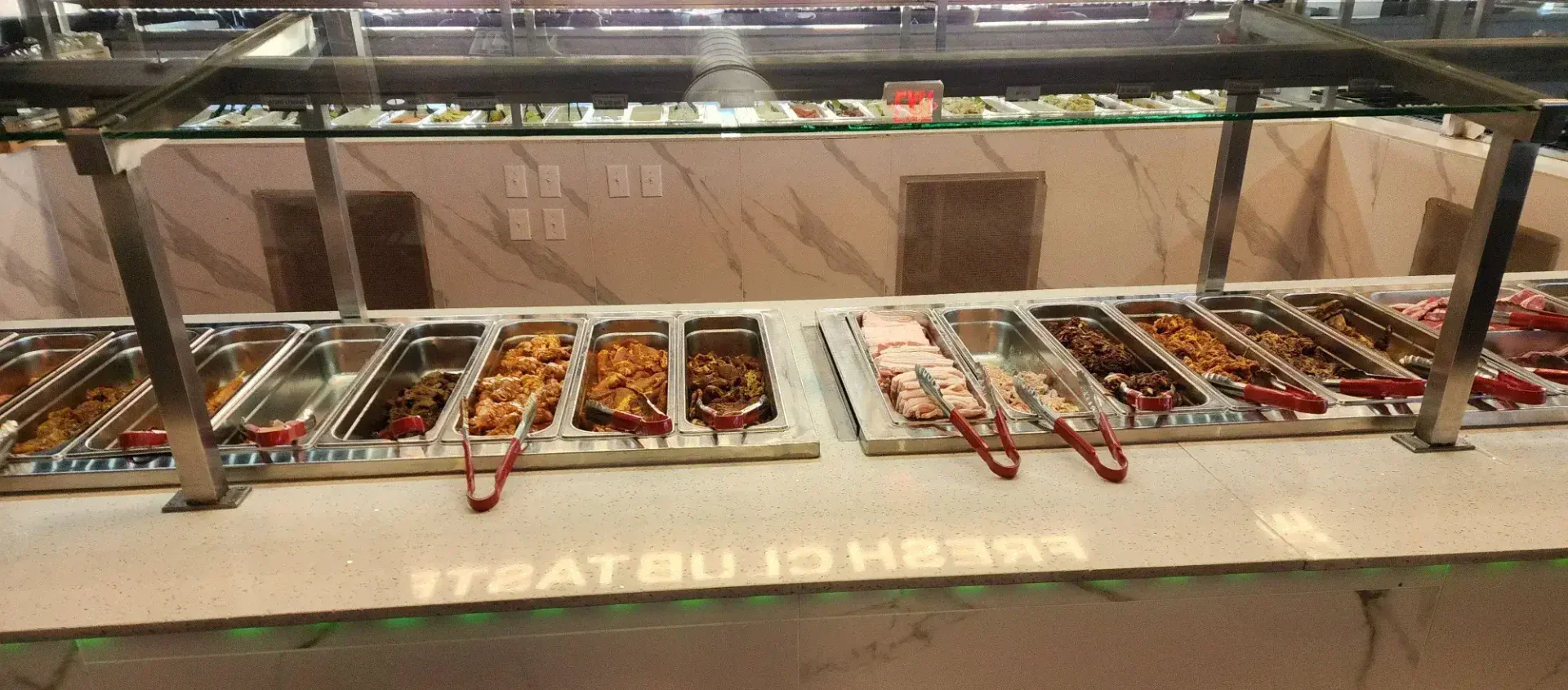 Korean BBQ self-serve buffet station with marinated beef, pork belly, and various meat selections in stainless steel trays